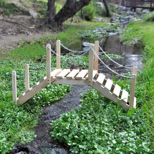 A small 3-foot wooden garden bridge with handrails and a simple slatted design spans a narrow stream, surrounded by lush green plants. It features rope chains connecting to vertical posts for added detail.