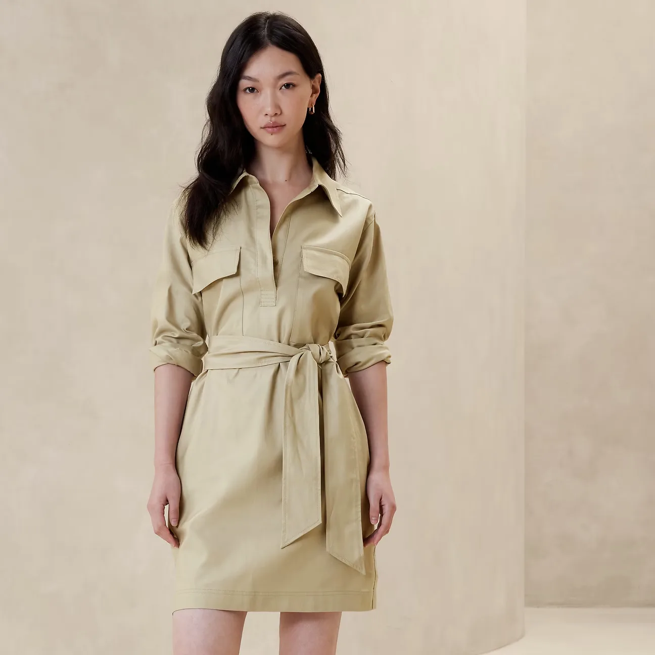 A woman models a knee-length, olive green satin shirt dress with rolled sleeves and a tie waist.