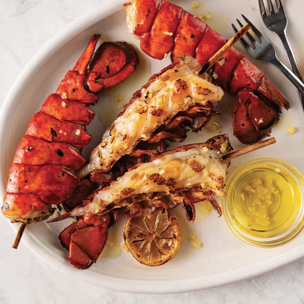Grilled lobster tails on skewers are served on a plate with lemon slices and a small dish of butter or oil, accompanied by two forks.