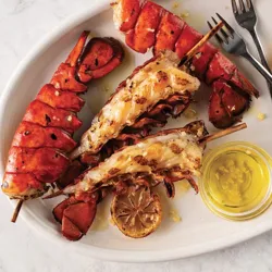 Grilled lobster tails on skewers are served on a plate with lemon slices and a small dish of butter or oil, accompanied by two forks.