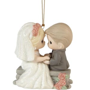 Two Precious Moments figurines depict a bride and groom sitting closely, holding hands. The bride wears a glittery white dress with a veil adorned with pink roses, while the groom is in a gray suit.