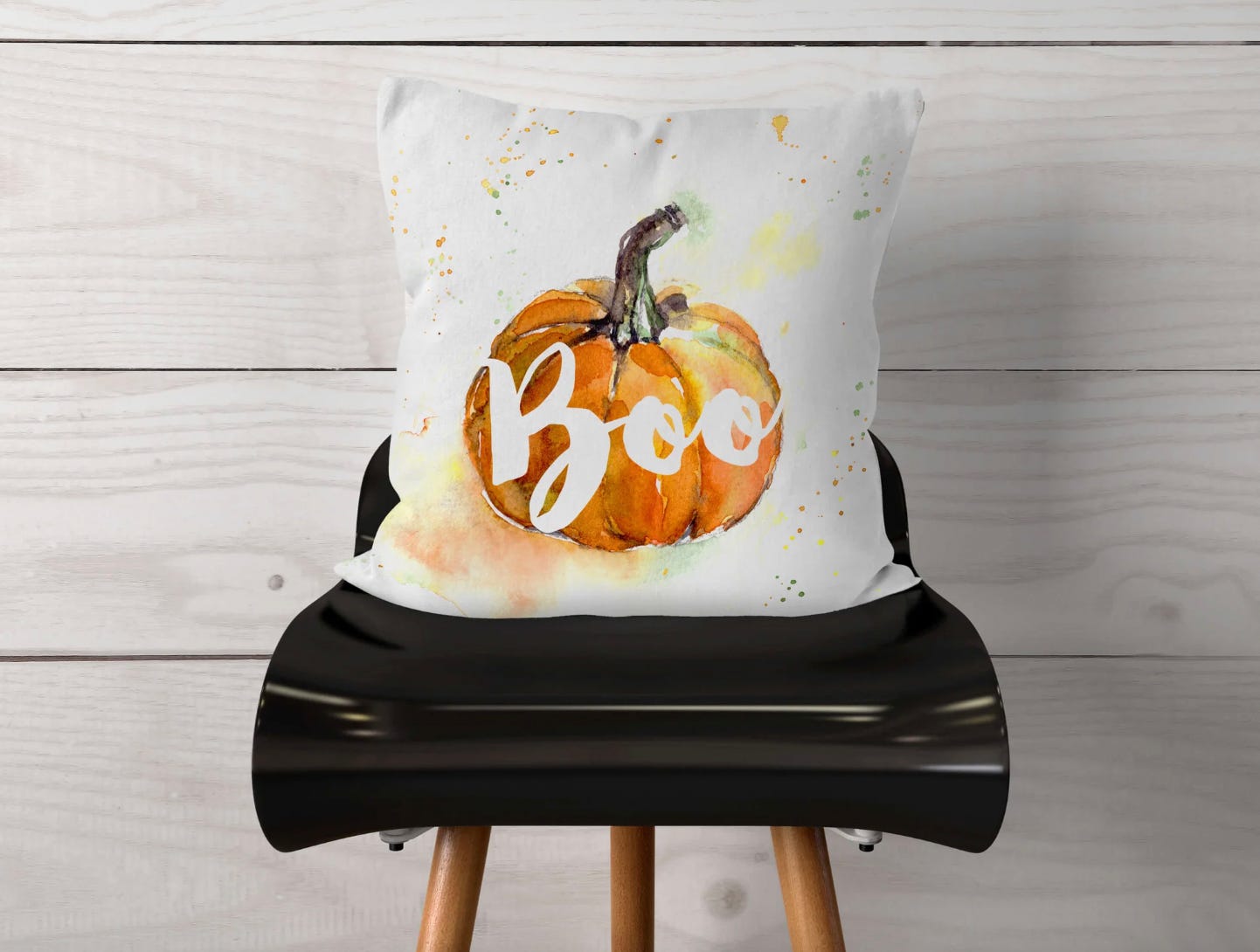 A square pillow cover with a watercolor painting of an orange pumpkin and the word \“Boo\“ in cursive script, displayed on a black chair against a white floor and wall backdrop.