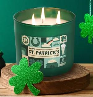 A green St. Patrick's Day-themed candle with three wicks, featuring holiday symbols like shamrocks and a horsehoe, is adorned with a glittery green shamrock decoration on a wooden stand.