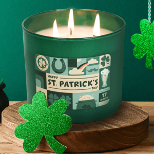A green St. Patrick's Day-themed candle with three wicks, featuring holiday symbols like shamrocks and a horsehoe, is adorned with a glittery green shamrock decoration on a wooden stand.