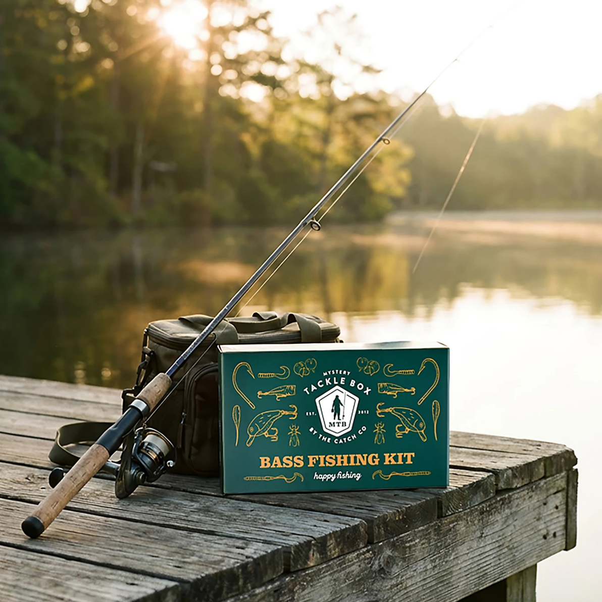 A fishing rod and bag sit next to a Mystery Tackle Box Bass Fishing Kit on a wooden dock, with a serene lake and sunlight in the background.