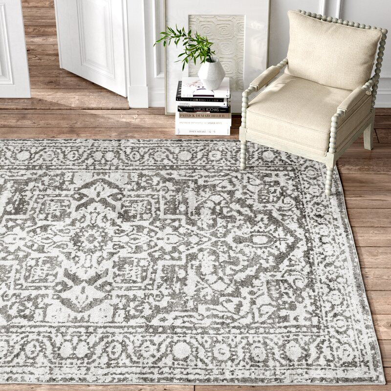 A traditional-style area rug with a distressed grey and white pattern, displayed in a room with a cream chair and a small side table.