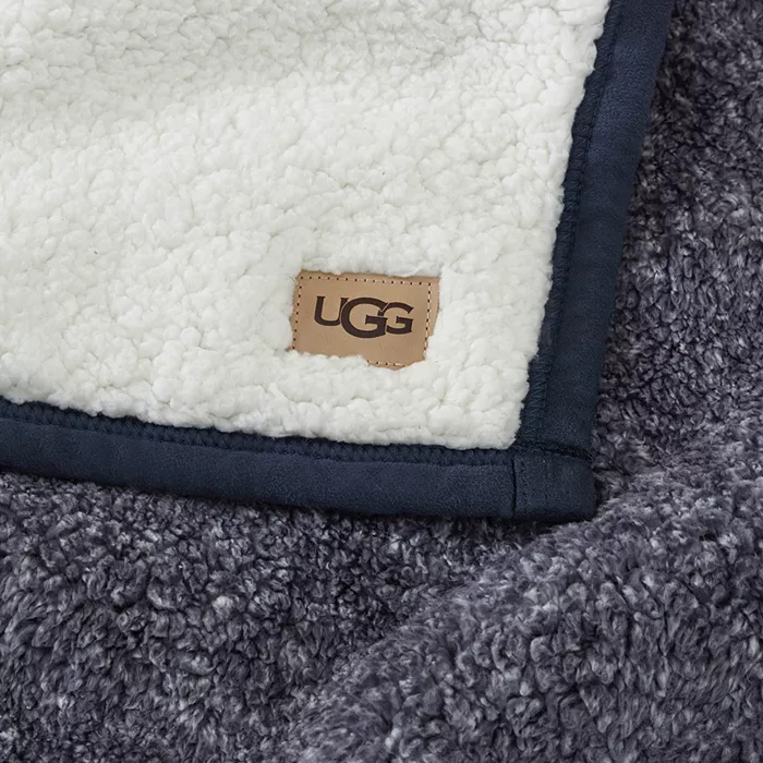 A dual-textured UGG throw blanket with a creamy white shearling side and a contrasting darker gray plush side, featuring a small rectangular UGG brand label.