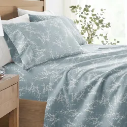 A bed with blue sheets featuring a white floral pattern, accompanied by white pillows. Nearby, a wooden side table holds a white lamp, a small succulent, and a clock. A leafy plant is positioned by the window, adding a touch of greenery.