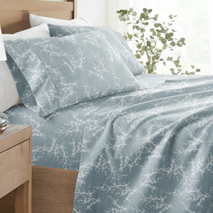 A bed with blue sheets featuring a white floral pattern, accompanied by white pillows. Nearby, a wooden side table holds a white lamp, a small succulent, and a clock. A leafy plant is positioned by the window, adding a touch of greenery.