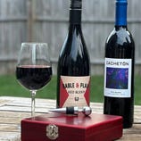 Two bottles of red wine, \“Gable & Plain\“ and \“Cachetón,\“ are on a table beside a filled wine glass and a red box with a wine stopper.