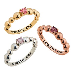 Three beaded rings in gold, silver, and rose gold, each featuring a pink gemstone and engraved with \“Charlotte Anne.\“