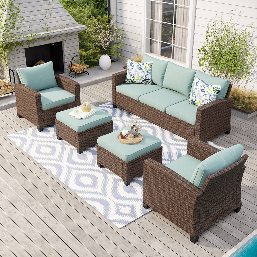 Outdoor patio set with a wicker design, featuring a three-seater sofa, two armchairs, and two ottomans, all with light blue cushions. An outdoor rug with a geometric pattern and decorative pillows add to the setup.