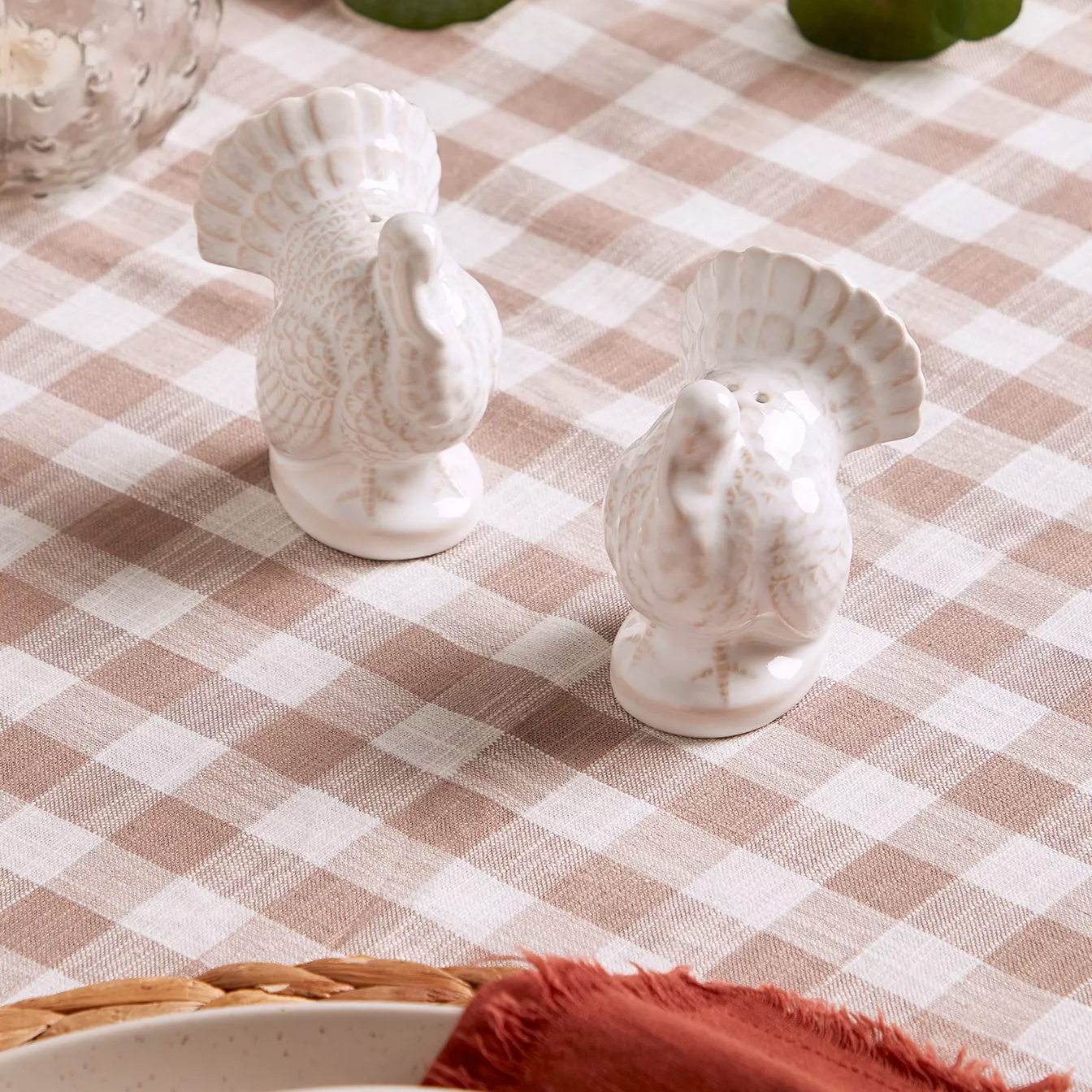 Two ceramic turkey-shaped salt and pepper shakers are displayed on a beige and white checkered tablecloth, accompanied by red cloth napkins and a woven basket.