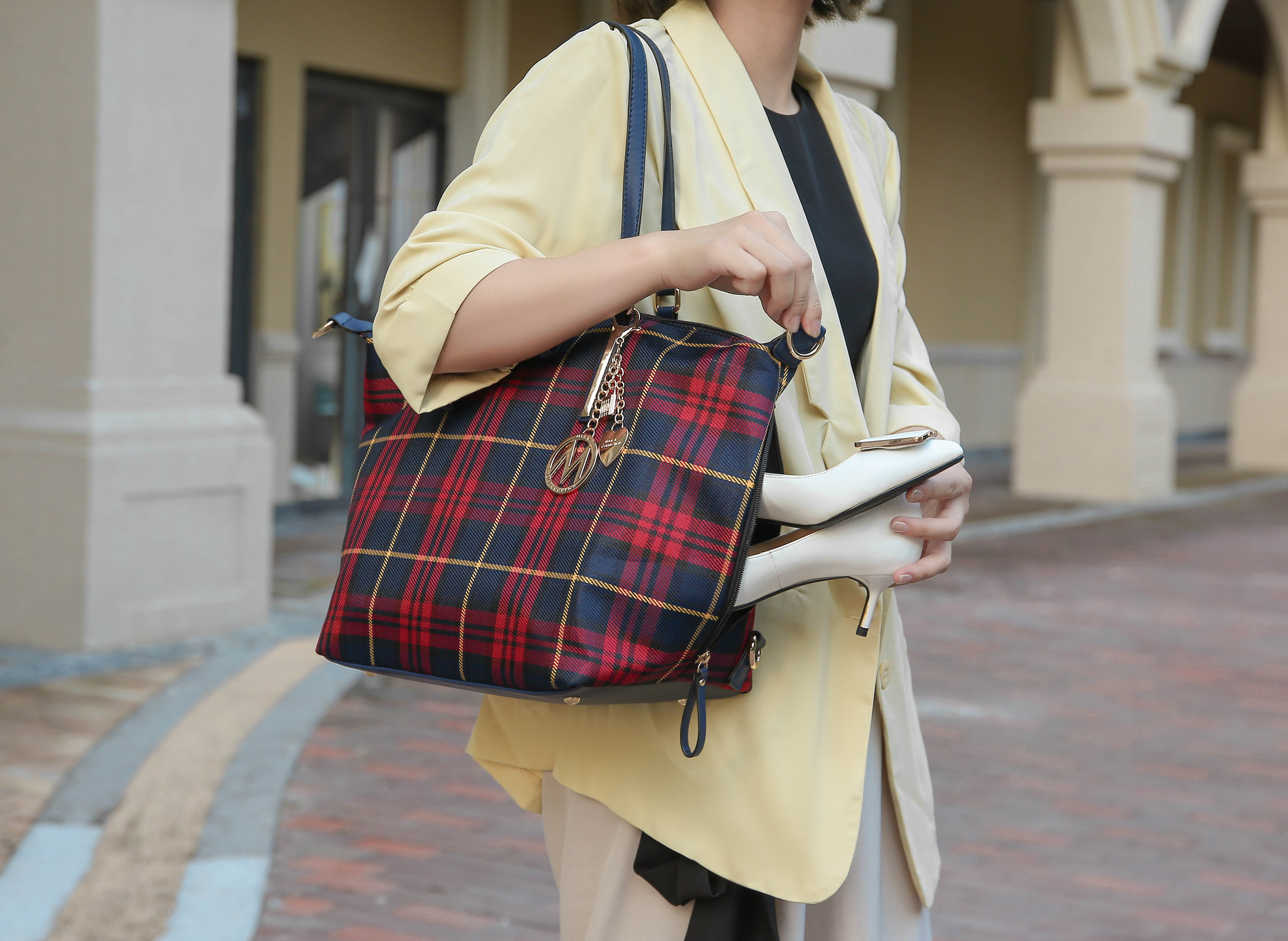 A person holding a plaid patterned tote bag and white high-heeled shoes.