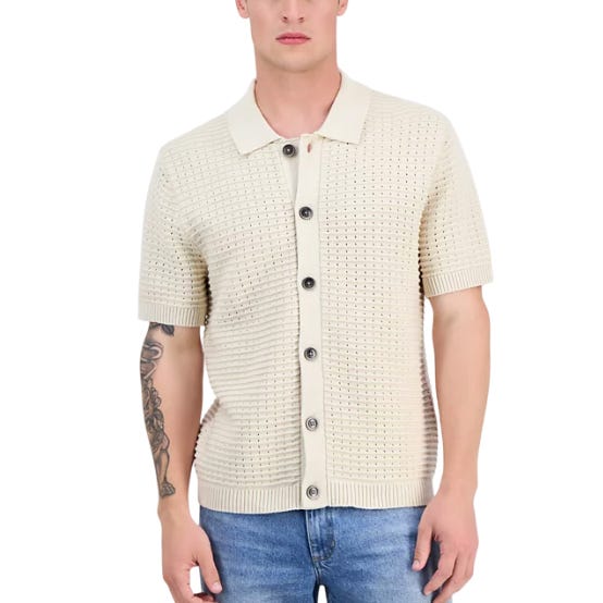 A man is wearing a cream-colored, short-sleeve, button-up cardigan with a textured, waffle-knit pattern. He also has a tattoo on his left arm and is wearing blue jeans.
