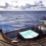 A cruise ship deck features a swimming pool surrounded by lounge chairs, overlooking the ocean under a cloudy sky.