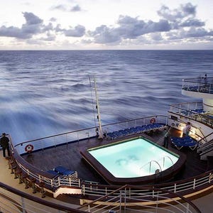 A cruise ship deck features a swimming pool surrounded by lounge chairs, overlooking the ocean under a cloudy sky.