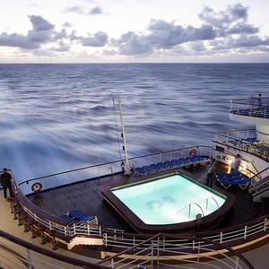 A cruise ship deck features a swimming pool surrounded by lounge chairs, overlooking the ocean under a cloudy sky.