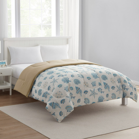 A bedroom features a white bed with a reversible comforter, displaying a beige underside and a blue seashell pattern on top. The room is minimalistic with white pillows, a nightstand, and a window.