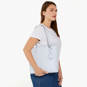 A woman wearing a white T-shirt and blue jeans is carrying a large light-colored shoulder bag.
