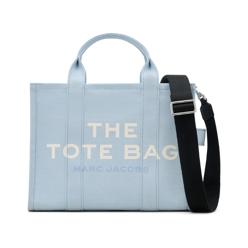 A light blue Marc Jacobs medium tote bag with a removable black strap, featuring the brand's name and \“THE TOTE BAG\“ text on the front.