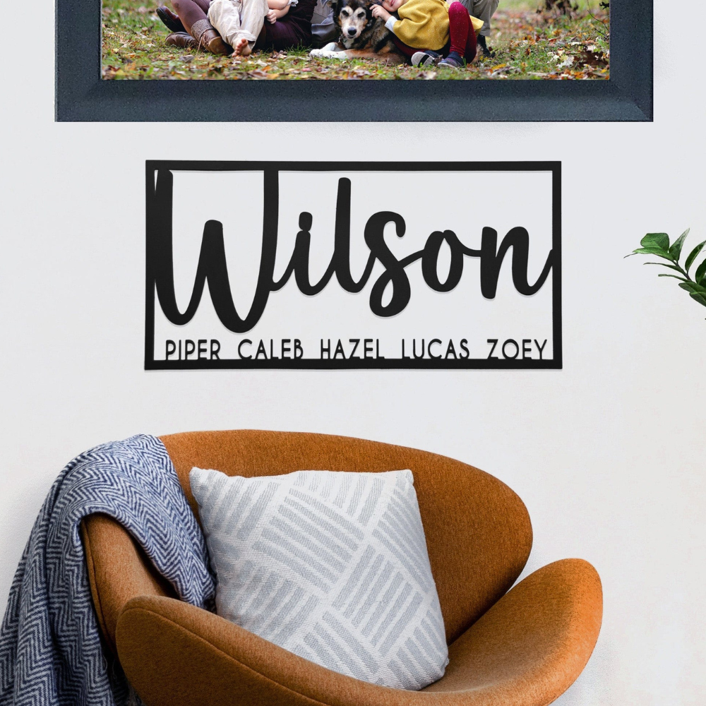A personalized wall sign displays the name \“Wilson\“ with the names Piper, Caleb, Hazel, Lucas, and Zoey beneath it. It is shown above a brown chair with a patterned pillow and a blue-and-white throw blanket.