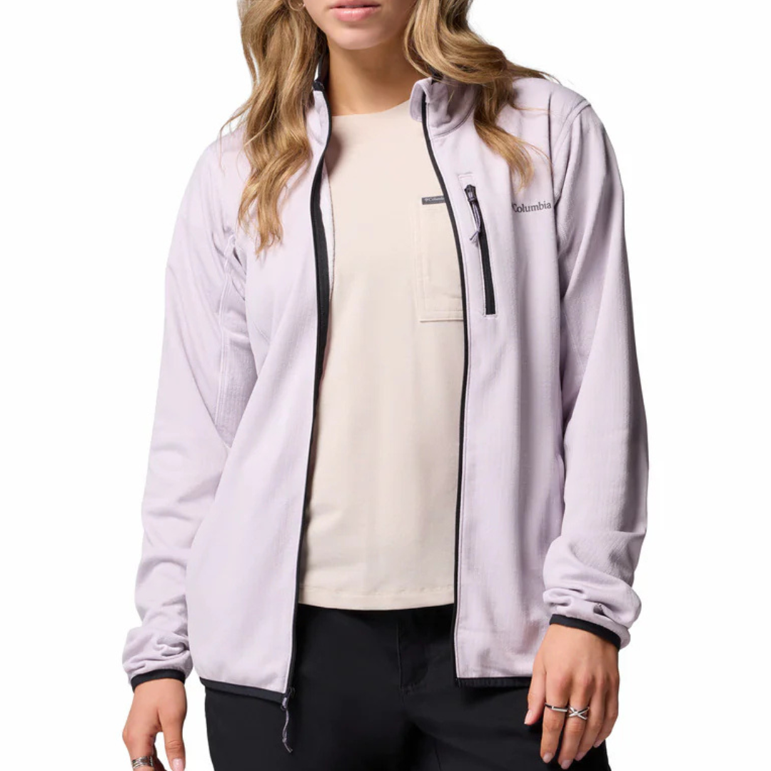 A woman is wearing a light-colored Columbia jacket with a zippered chest pocket and black trim, paired with a cream t-shirt and dark pants.