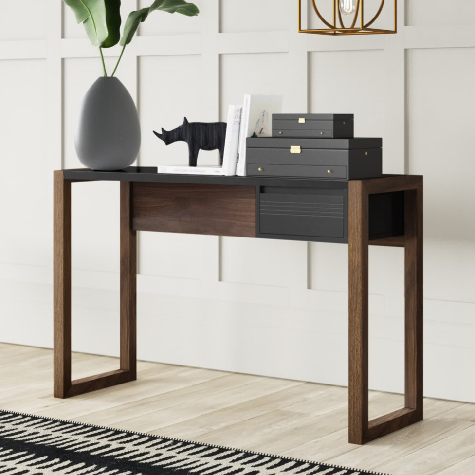 A modern console table with wooden legs and a black top, adorned with a large vase with green leaves, a decorative rhinoceros figure, stacked black boxes, and a few books, set against a white paneled wall with a geometric pendant light above.