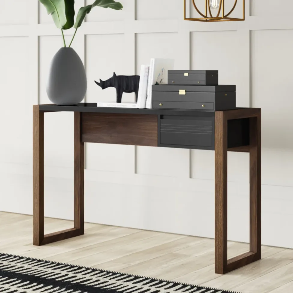 A modern console table with wooden legs and a black top, adorned with a large vase with green leaves, a decorative rhinoceros figure, stacked black boxes, and a few books, set against a white paneled wall with a geometric pendant light above.