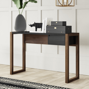 A modern console table with wooden legs and a black top, adorned with a large vase with green leaves, a decorative rhinoceros figure, stacked black boxes, and a few books, set against a white paneled wall with a geometric pendant light above.