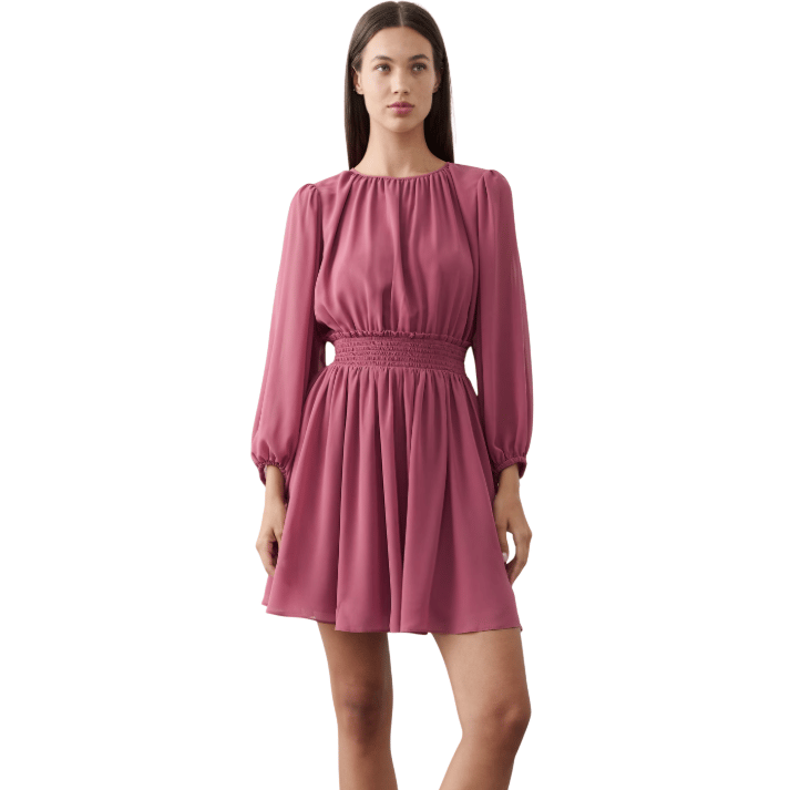A woman is wearing a mauve, long-sleeve mini dress with a gathered neckline, cinched elastic waist, and flowy skirt, paired with black flat shoes.