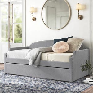 The Kelly Clarkson Jordane Daybed features a gray upholstered frame with armrests, nailhead trim, and a pull-out trundle. It is complemented by a round mirror, wall sconces, and a patterned blue-gray rug.