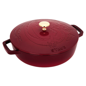 Staub Cast Iron Braiser with a dragon-embossed lid and gold knob, in a deep red color. The handles are wide for easy gripping, and the cookware features sturdy construction for even heat distribution.