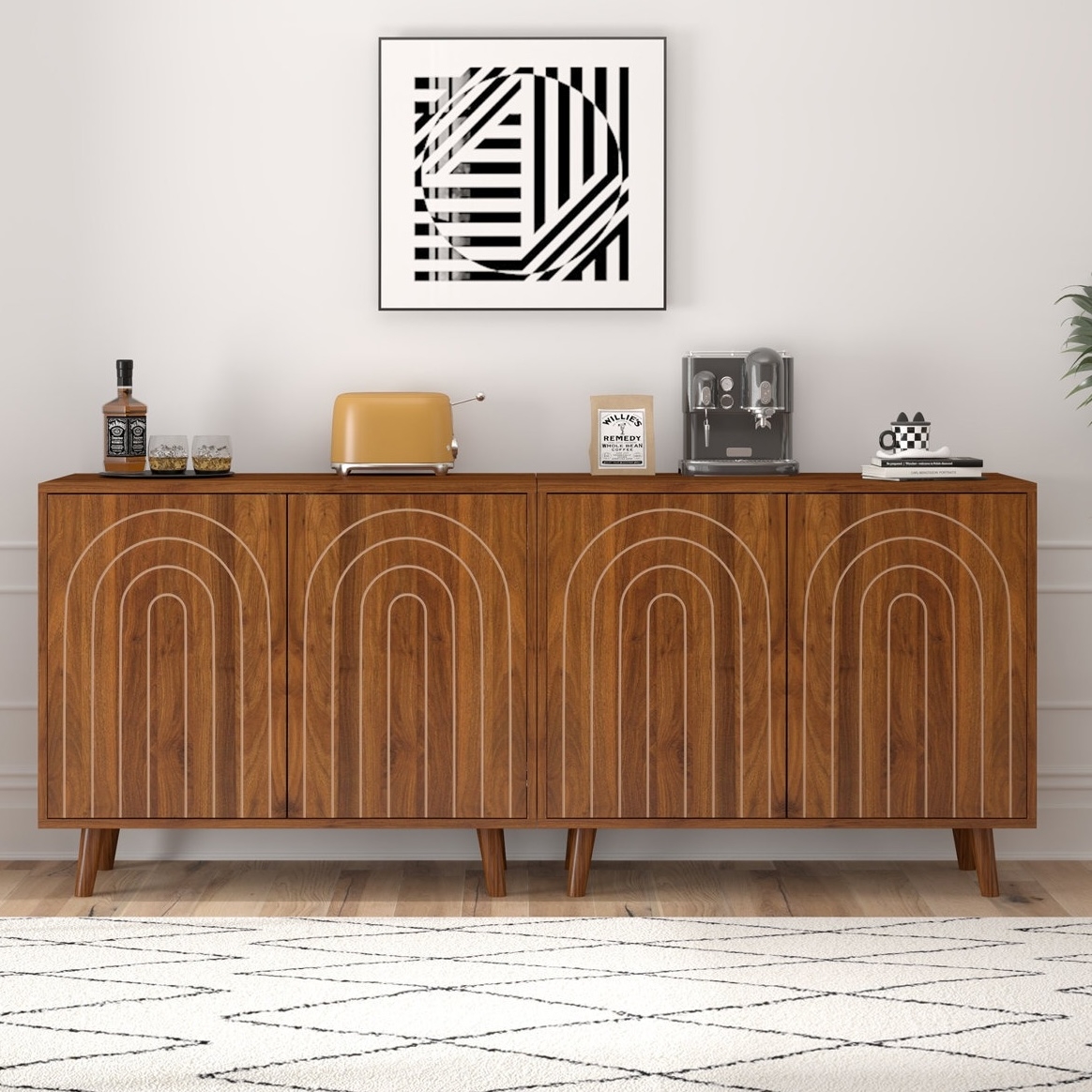 A mid-century modern wooden sideboard with white geometric patterns, topped with a whiskey bottle, glasses, an orange toaster, coffee machine, and decor items. A black and white abstract art piece hangs above.