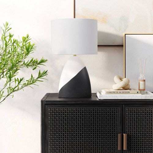 A modern table lamp with a dual-tone black and white base stands on a dark cabinet. Nearby are decorative items and a plant, creating a stylish interior setting.