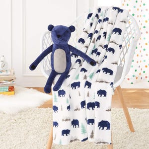 A stuffed blue bear and a white blanket with blue and green bear and tree patterns draped over a white chair.