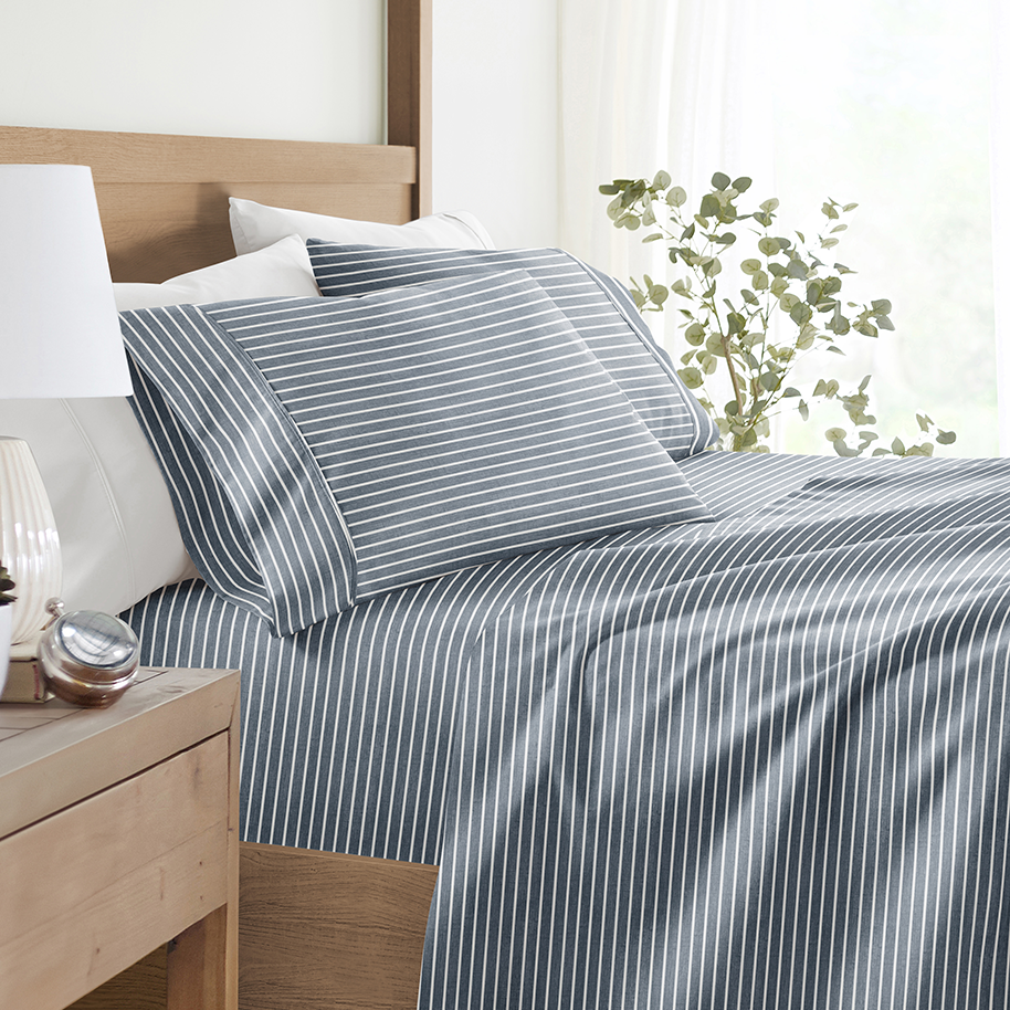 A bed features blue and white striped bedding with matching pillowcases. A wooden nightstand holds a lamp and decorative items, and a plant is visible in the background.