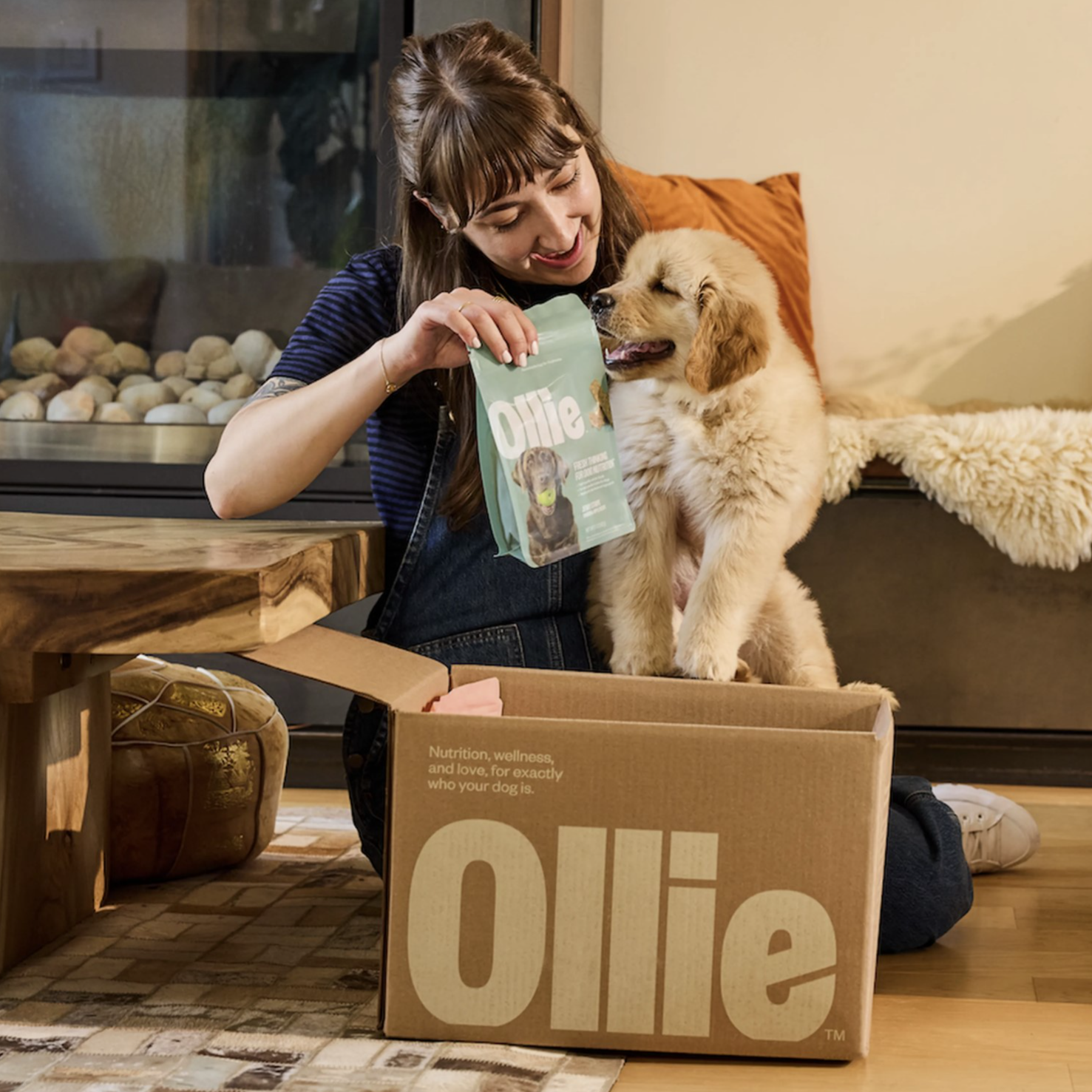 A woman is holding a bag of Ollie dog food next to a fluffy puppy. They are sitting by a fireplace with a box labeled \“Ollie\“ nearby.