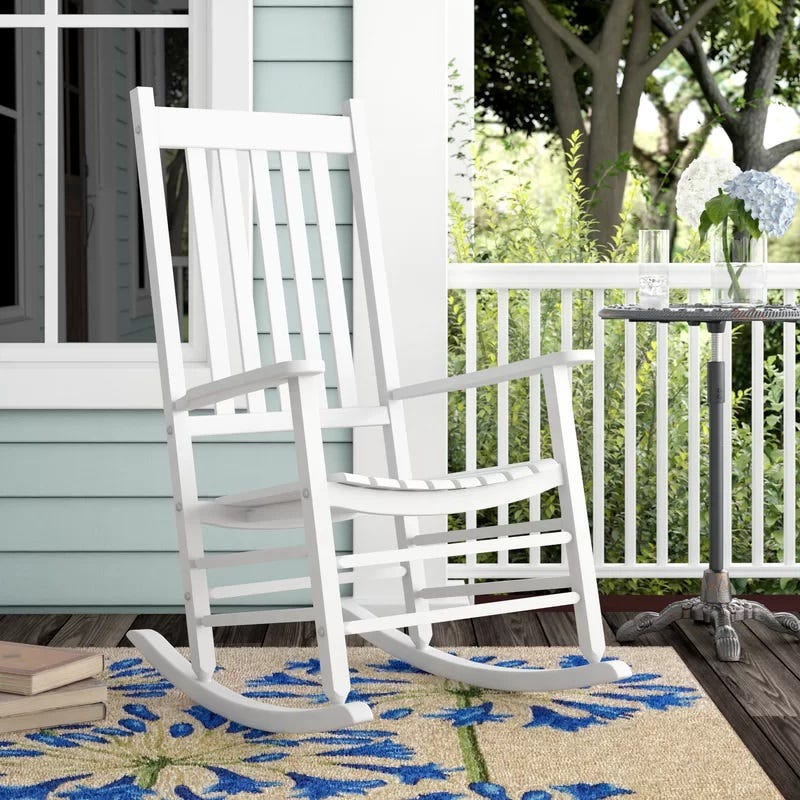 A white wooden rocking chair is positioned on a porch next to a small table with a glass and a vase with flowers.
