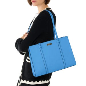 A woman holds a bright blue Kate Spade large tote with two shoulder straps and a structured rectangular shape. The bag features a small logo plate on the front and is worn over a black and white cardigan.
