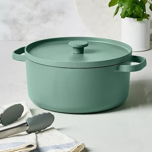 A green cast iron dutch oven with a matching lid and side handles, placed next to metal tongs and a striped cloth.
