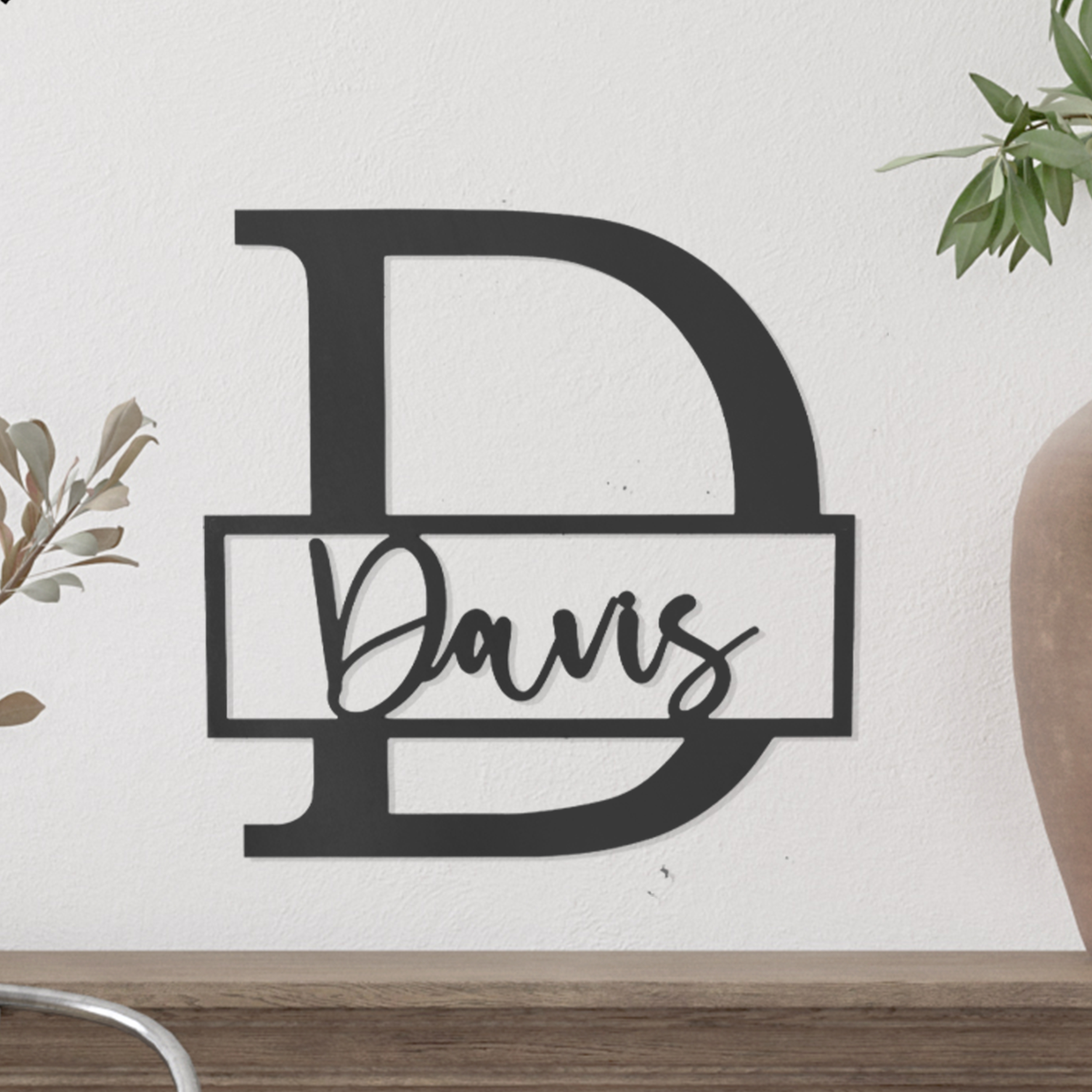 A black metal wall decor featuring a large letter \“D\“ with the name \“Davis\“ in a script font across the center, set against a white wall with plants in the background.