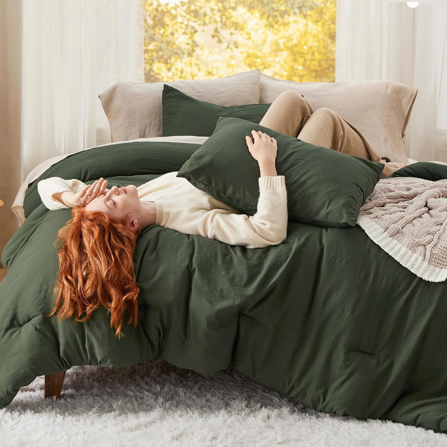 A person lies relaxed on a bed with dark green bedding, surrounded by pillows and a neutral knit blanket, set in a bright room with large windows.