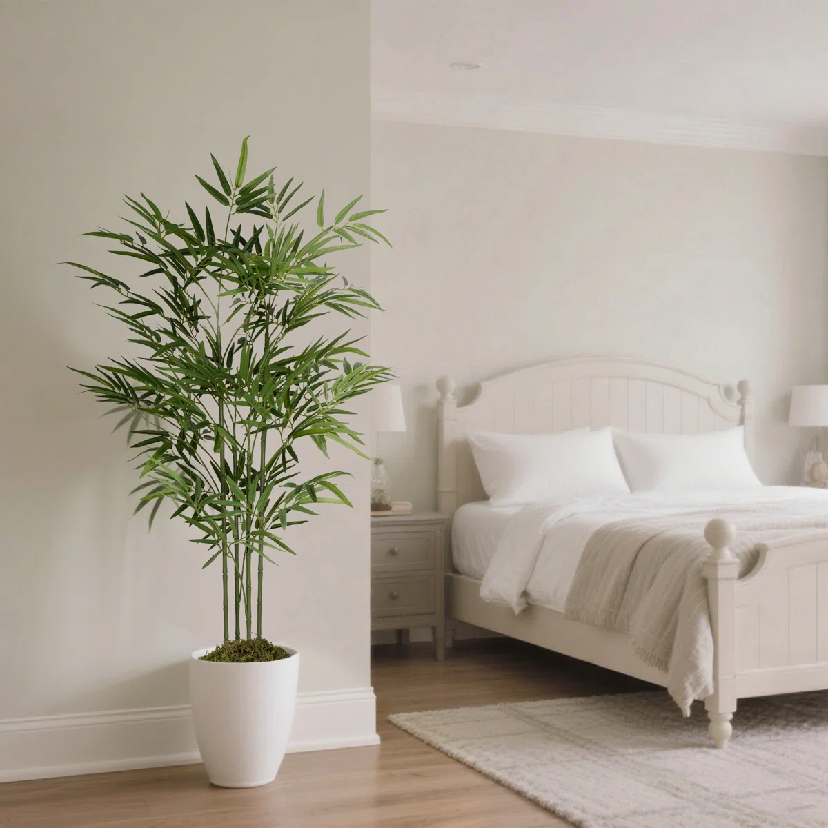 A potted artificial bamboo plant is in a white pot placed in a minimalist, neutral-toned bedroom with a white bed, side tables, and two lamps.