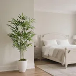 A potted artificial bamboo plant is in a white pot placed in a minimalist, neutral-toned bedroom with a white bed, side tables, and two lamps.