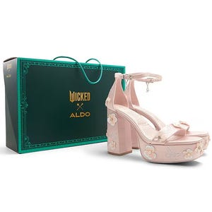 The Aldo X Wicked Platform Sandal is a pink high-heeled shoe adorned with floral and butterfly designs. It features an ankle strap and comes in a green Wicked-branded box.