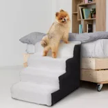 A Pomeranian dog stands on a plush, three-step pet staircase, designed to help small pets access higher places like beds or couches. The stairs are covered in a soft, white material with black sides.