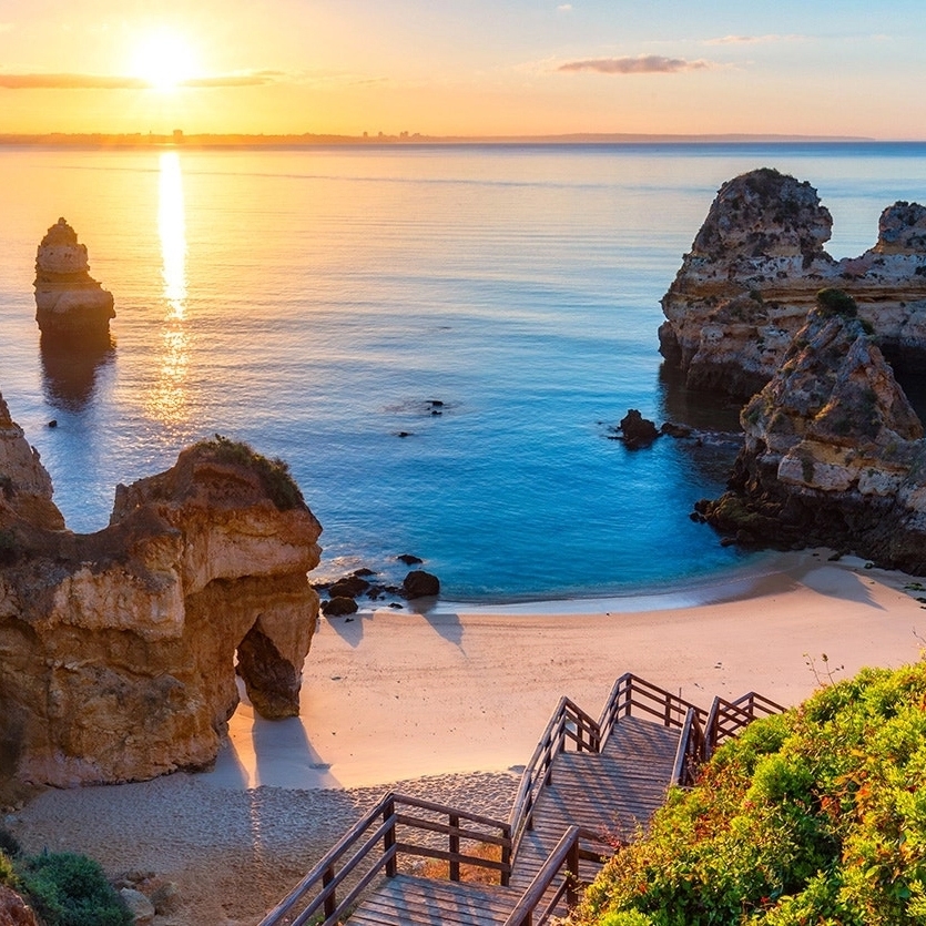A tranquil beach scene at sunset with rock formations, a calm sea, and wooden stairs leading down to the sand.