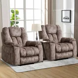 Two plush brown recliner chairs with padding and armrests, separated by a small table holding a lamp and a teacup, are placed on a light-colored rug in a well-lit room with large windows.