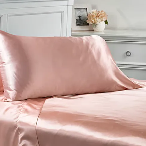 A pink satin pillowcase and matching bedsheet set on a bed, with a white headboard and a grey dresser in the background, decorated with a vase of flowers.